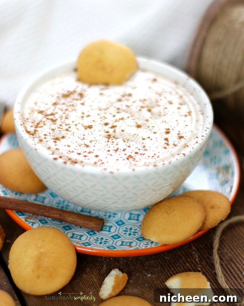 Eggnog dip in a bowl on a plate surrounded by vanilla wafer cookies and a cinnamon stick.